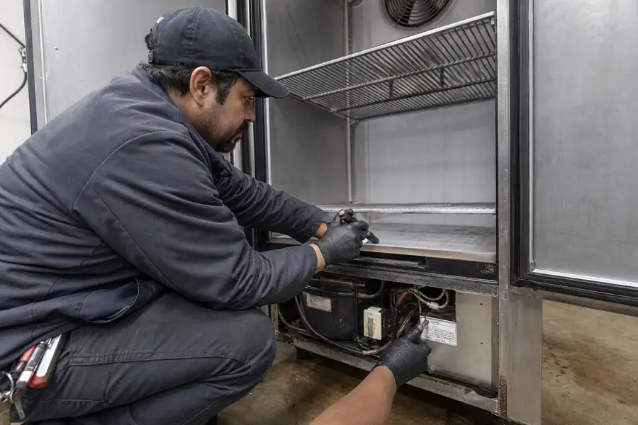 Action-oriented service photograph for “Where to Find the Data Plate on Commercial Refrigeration, Brand by Brand.” Show an experienced commercial refrigeration gasket technician carrying out the central inspection, measurement or service step described in “Where to Find the Data Plate on Commercial Refrigeration, Brand by Brand”. Include technician hands and only the appropriate practical tools where relevant, with the commercial refrigeration door or seal clearly visible and the process easy to understand from the image alone. Relevant equipment: Reach-ins; walk-ins; prep tables; merchandisers; undercounter units. Relevant manufacturers when naturally visible: True, Traulsen, Turbo Air, Beverage-Air, Kolpak, Nor-Lake, Delfield, Continental, Hoshizaki, Master-Bilt. Authentic field-service atmosphere, realistic equipment condition, no promotional posing, 16:9 landscape.