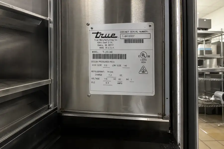 Editorial hero photograph for a commercial foodservice refrigeration article titled “Where to Find the Data Plate on Commercial Refrigeration, Brand by Brand.” Show One data plate photo per brand covered, shot in place with the surrounding door visible for orientation. Relevant equipment: Reach-ins; walk-ins; prep tables; merchandisers; undercounter units. Relevant manufacturers when naturally visible: True, Traulsen, Turbo Air, Beverage-Air, Kolpak, Nor-Lake, Delfield, Continental, Hoshizaki, Master-Bilt. Use an authentic restaurant, grocery, school-kitchen or foodservice setting, natural practical lighting, realistic wear and materials, documentary composition, no staged corporate poses, no added text, 16:9 landscape. When the brief requires technical proof, publication should use a real job-site photo instead of an invented representation.