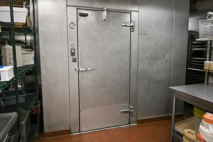 Editorial hero photograph for a commercial foodservice refrigeration article titled “Walk-In Cooler Door Problems and What Each One Is Telling You.” Show Annotated photo of a complete walk-in door. Relevant equipment: Walk-in coolers and freezers; swing doors; sliding doors; site-built doors. Relevant manufacturers when naturally visible: Kolpak, Nor-Lake, Master-Bilt, Bally. Use an authentic restaurant, grocery, school-kitchen or foodservice setting, natural practical lighting, realistic wear and materials, documentary composition, no staged corporate poses, no added text, 16:9 landscape. When the brief requires technical proof, publication should use a real job-site photo instead of an invented representation.